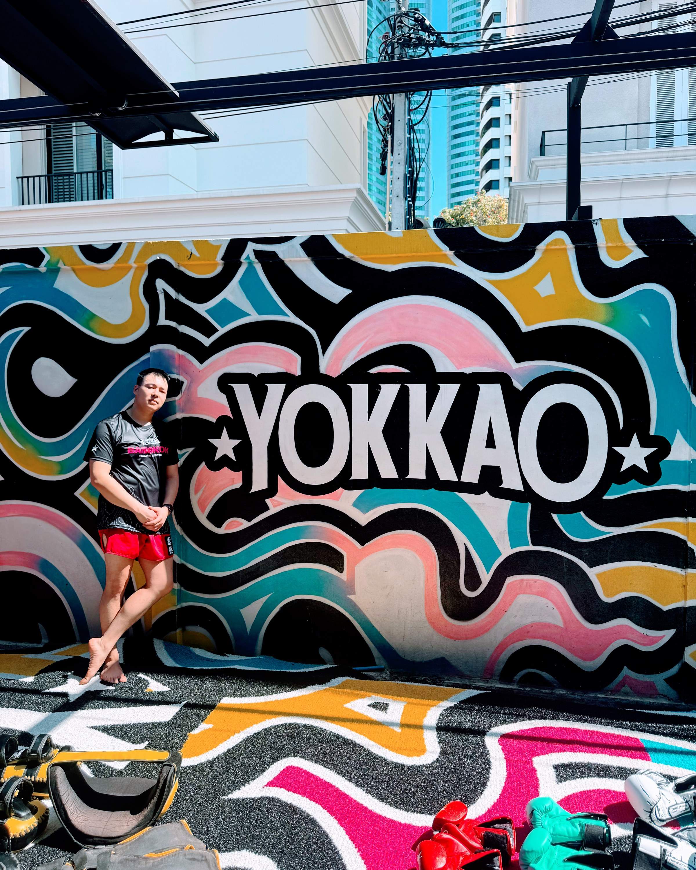 Standing in front of a colorful mural featuring the word "YOKKAO", muay thai gear lying on the ground under the sun