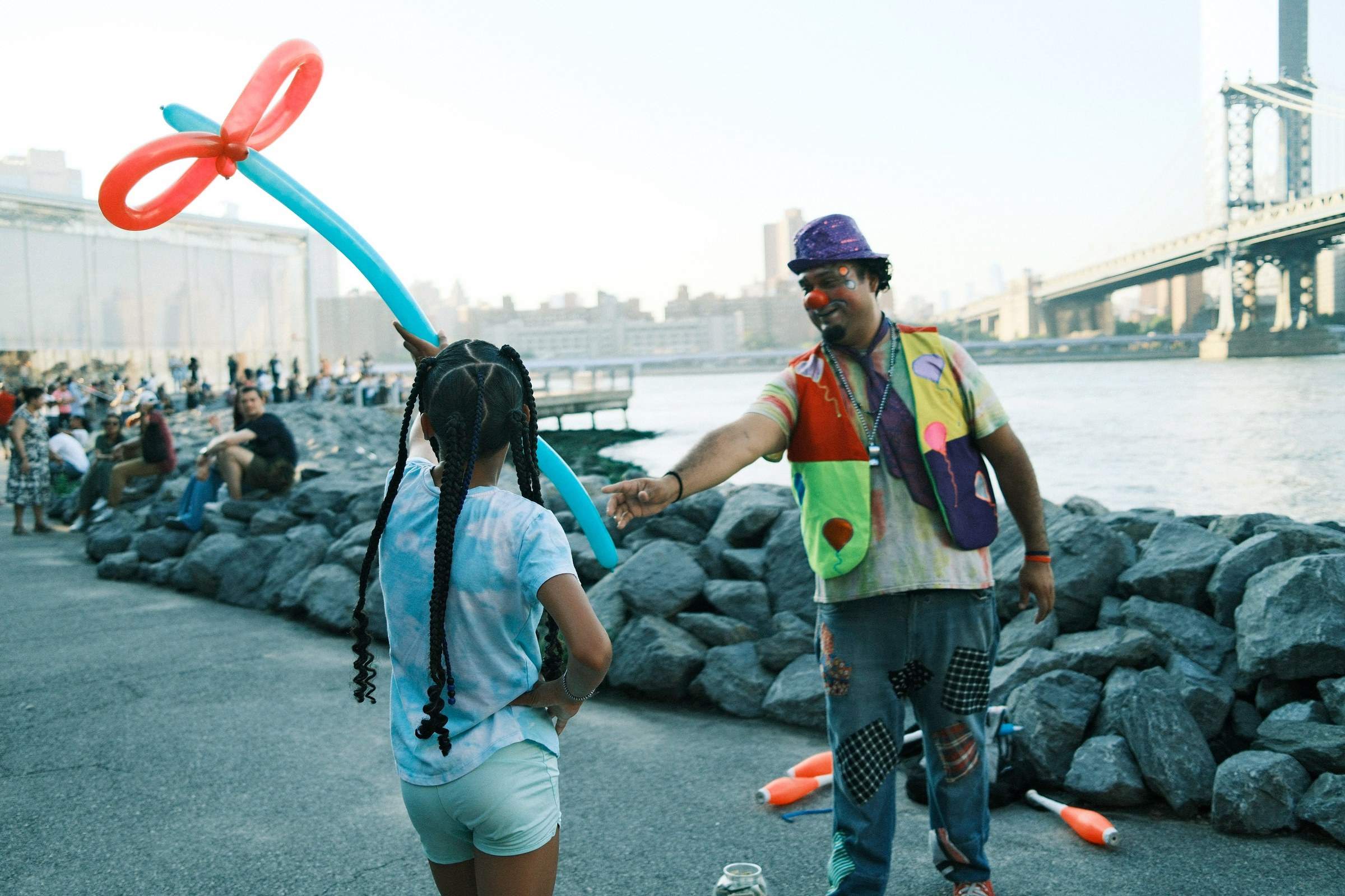 A colorful performer in a vibrant outfit and purple hat presents a large balloon animal to a child with braided hair on the DUMBO riverbank, with a crowd gathered and a bridge in the hazy skyline.