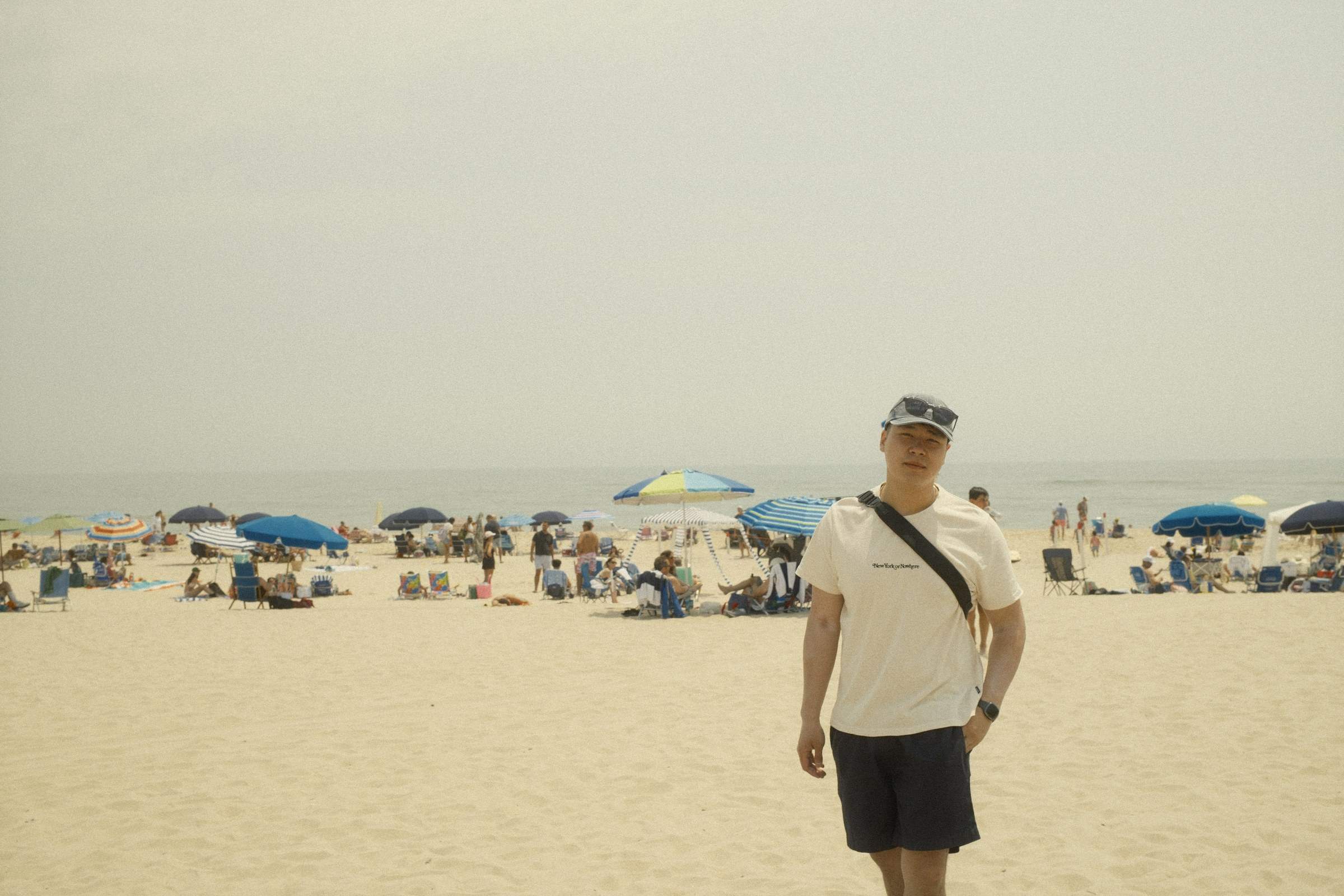 Derek in a white shirt and black shorts, standing on a sandy beach in the Hamptons, surrounded by colorful umbrellas and beachgoers, with a clear sky and ocean in the background.