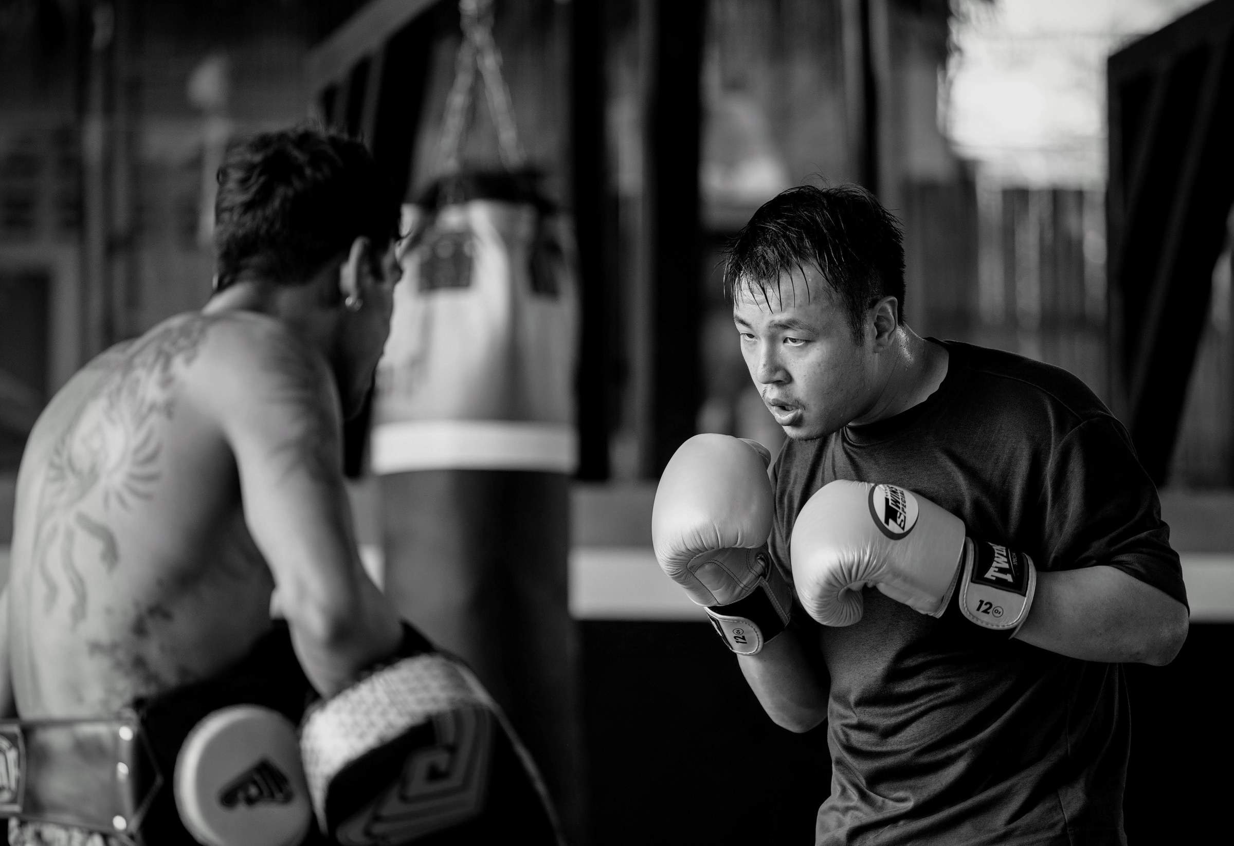 Black and white photo of Derek training at Punch It gym with a Kru, with muay thai pads, boxing gloves and a punching bag in view.