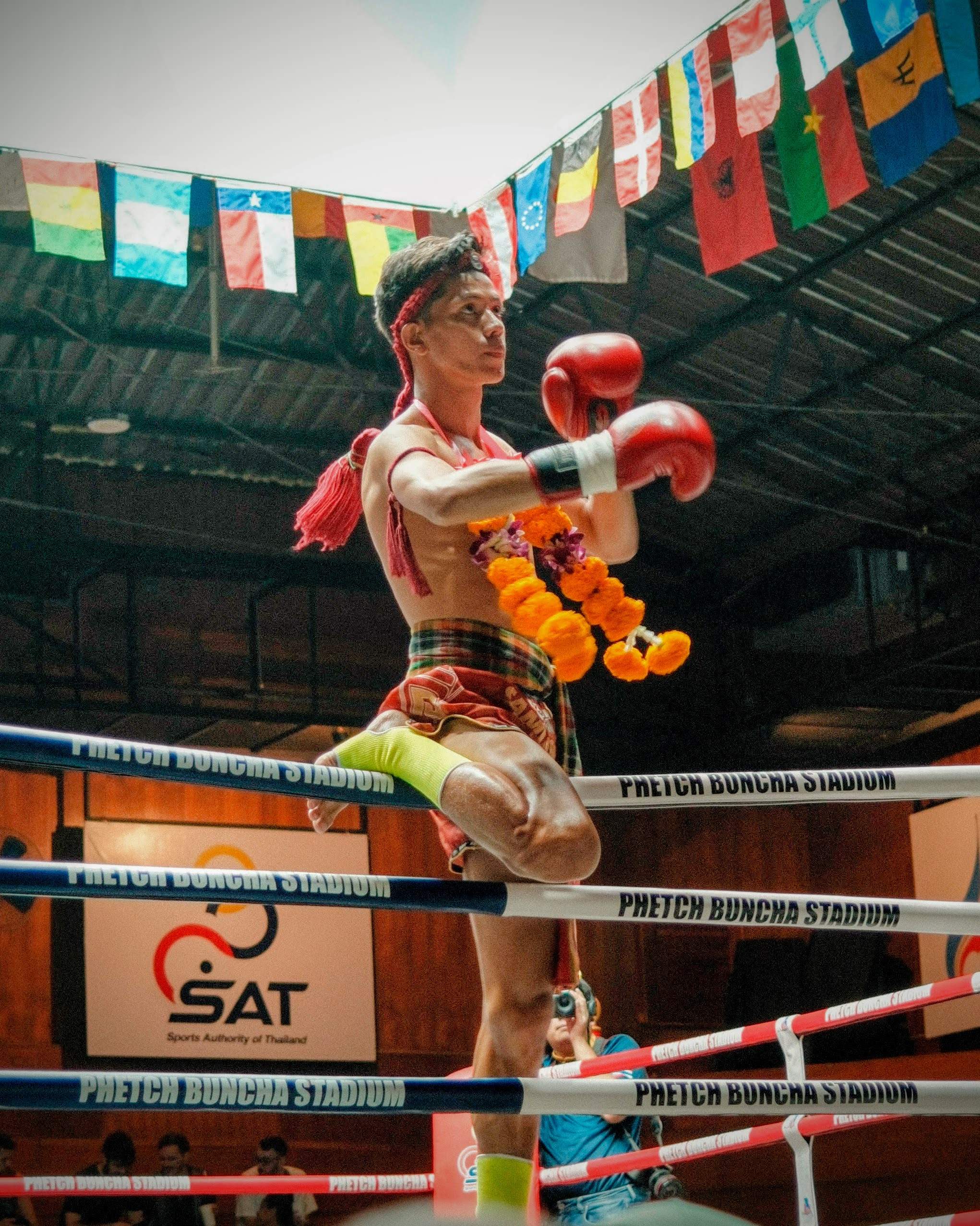 A Muay Thai fighter is performing a wai kru, a traditional pre-fight ritual, in a boxing ring adorned with a garland, under a canopy of international flags at Phetchbuncha Stadium.