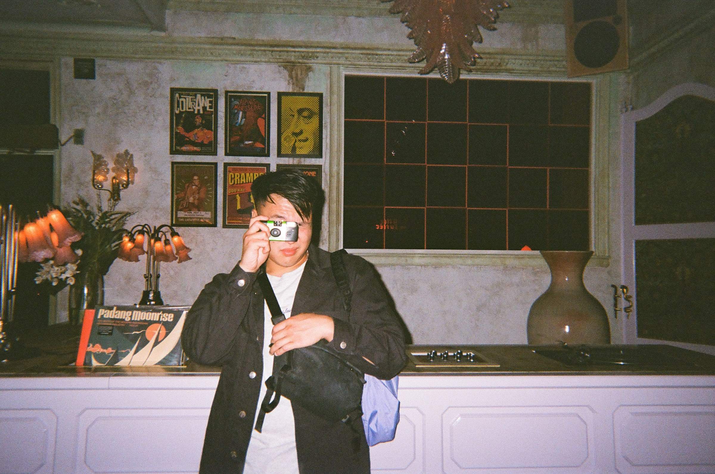Derek in front of a record and bar counter, facing the camera, taking a photo of the cameraperson with a disposable camera
