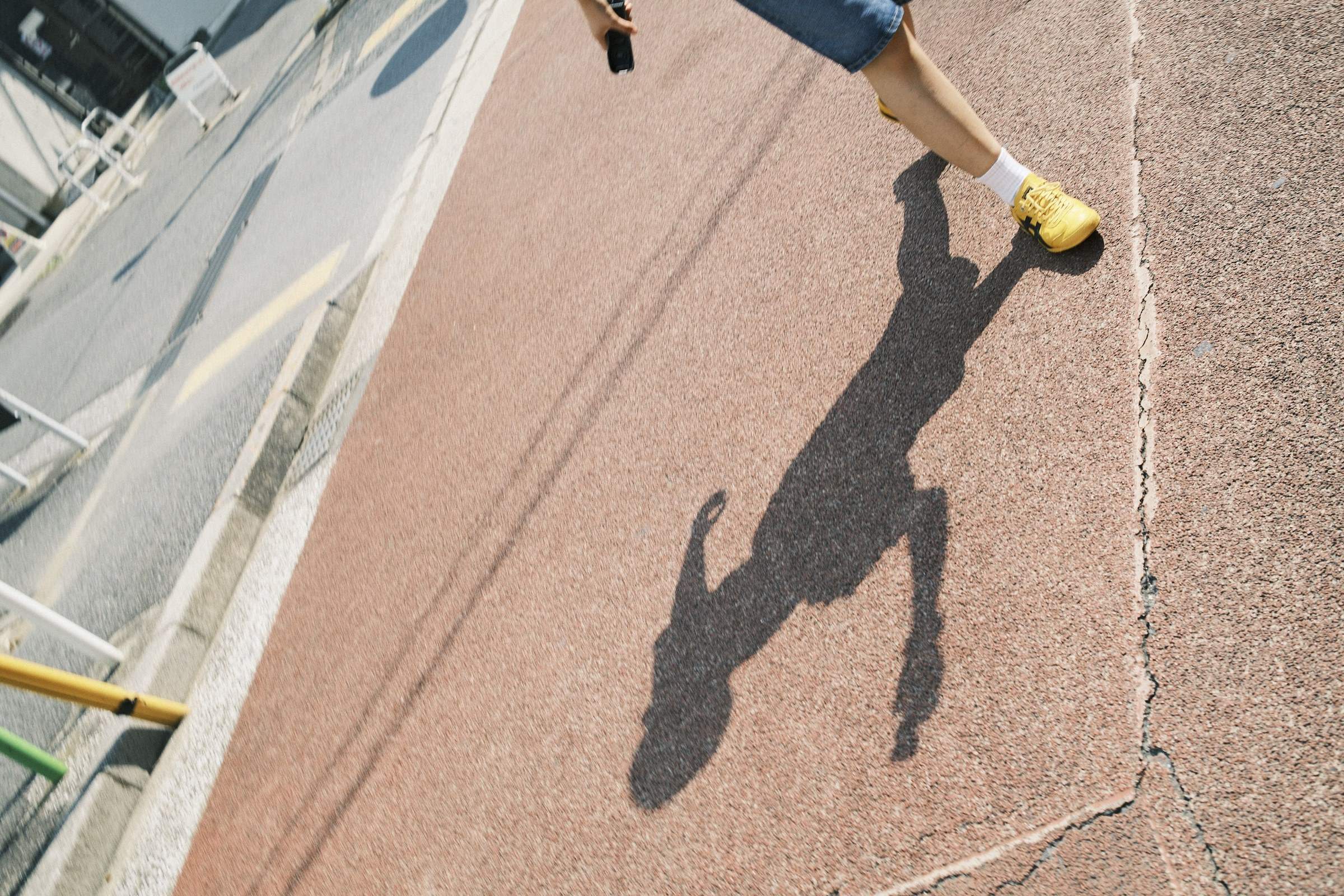 A person's shadow cast on a faded brick sidewalk, with the individual wearing shorts and yellow shoes, walking in sunlight.